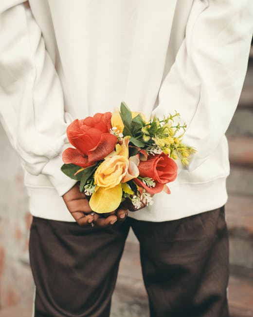 A person dressed in a white hoodie and dark pants is holding a floral bouquet behind their back. The bouquet features a combination of large, soft coral and yellow roses, complemented by smaller yello