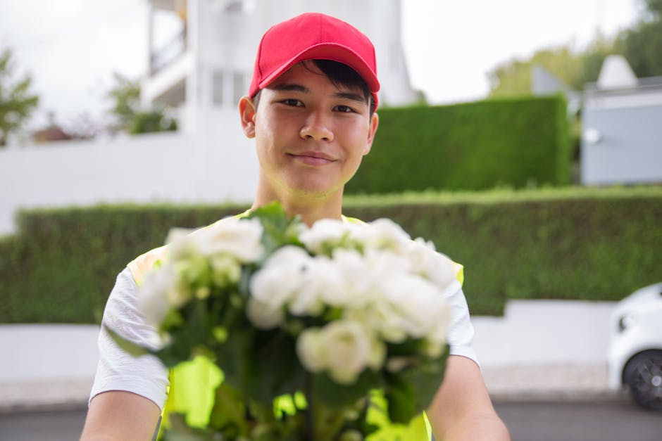 A young male flower delivery courier with a friendly expression, wearing a red cap and a high-visibility vest, is handing over a bouquet of fresh white flowers outdoors. The bouquet features a variety