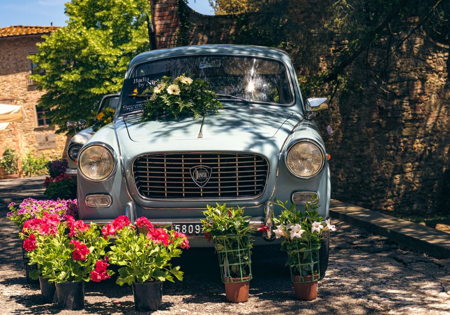 A vintage light blue car is parked outdoors in a sunny setting with lush green trees and historic stone buildings in the background. The car's hood features a lush floral arrangement consisting of whi