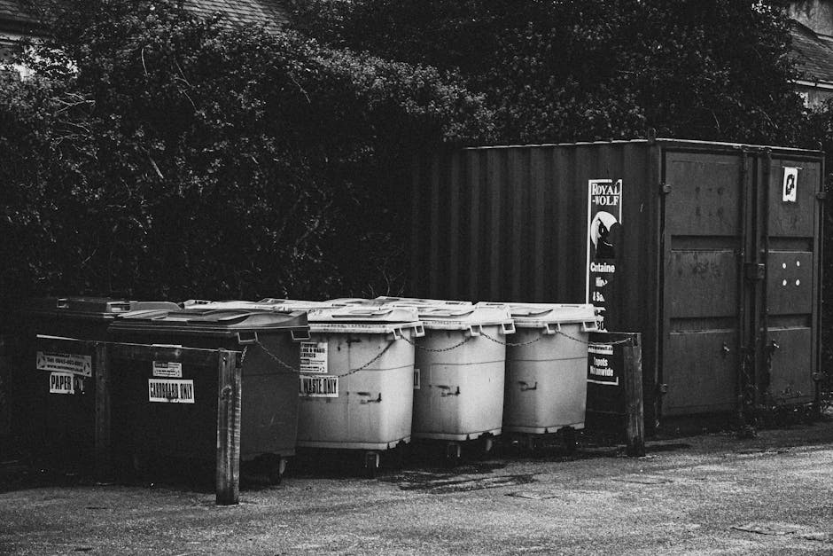 Black and white image showing a row of large white waste bins and a dark large container placed outdoors on a paved surface. The waste bins are labeled for recycling and waste disposal, with some lids