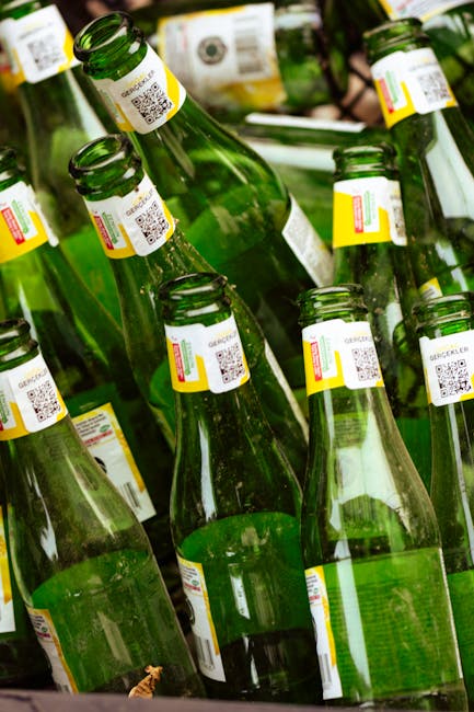 A collection of empty green glass bottles with attached QR code labels, arranged closely together, some standing upright while others are slightly tilted. The bottles are clear, showing reflections of