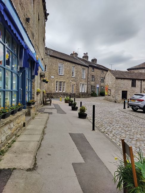 A narrow sidewalk along a historic stone building with blue window shutters and window boxes filled with small yellow flowers, leading into a cobblestone courtyard with parked cars and additional ston