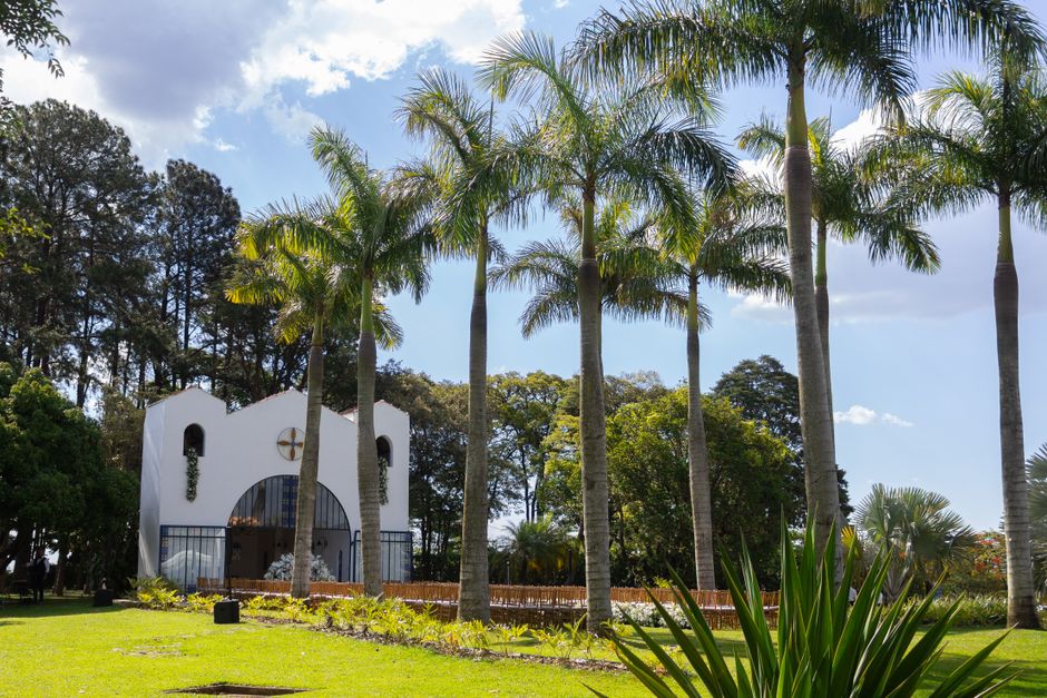 A white chapel with a curved roof and black wrought-iron balcony, set amidst tall palm trees and lush greenery under a partly cloudy sky, forming a scenic backdrop suitable for a wedding ceremony at V