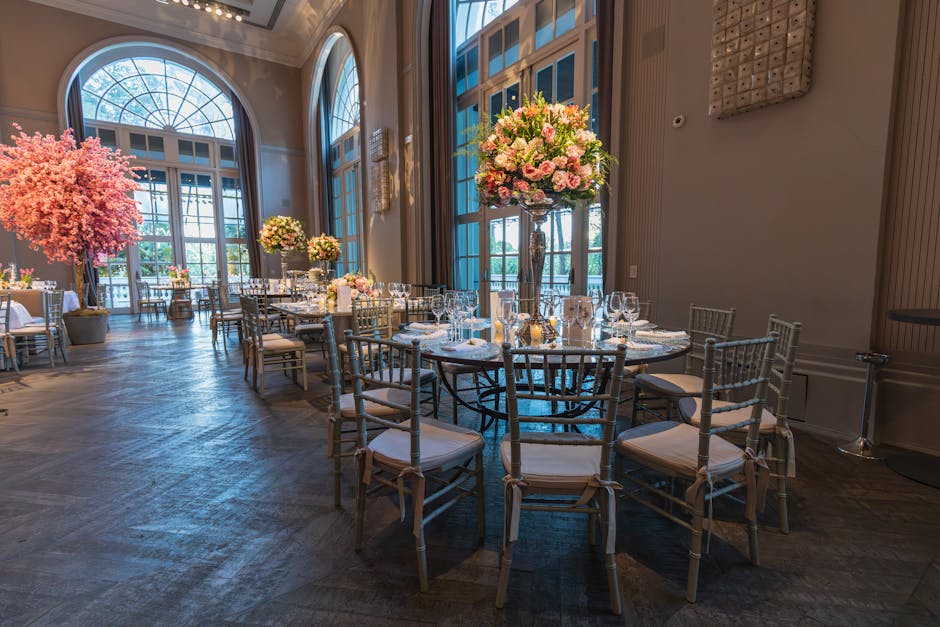 An elegantly decorated banquet hall featuring tall floral arrangements with pink, white, and blush roses, complemented by greenery, positioned on tall glass vases atop round tables set for an event. T