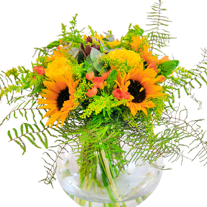 Upon first glance, this bouquet radiates sheer joy-swathes of golden sunflowers and opulent yellow roses burst forth, their brilliantly layered petals like little suns blooming over the urban gardens of Old Ford. The sunflowers' cocoa-brown hearts form a warm anchor amidst the bouquet, while rich yellow-orange rays fan out in a visual harmony that feels both lively and comforting. Interspersed within, yellow garden roses bloom with tightly nestled, spiral-shaped petals-textural marvels inviting touch. Glossy Hypericum berries, vivid red against the yellow, add sparkling accents as striking as Old Ford's canal boats in morning light. A diverse tapestry of greenery ties the whole together: fine fern fronds flow in graceful arcs, goldenrod sprigs with tiny yellow blossoms light up the gaps, and elongated eucalyptus leaves with their blue-green cast peek out, hinting at woodland walks through Tower Hamlets Cemetery Park. The arrangement sits nestled in a perfectly round, clear glass vase, its transparency revealing the lush stems and glimmering water-a symbol of freshness and honest craftsmanship from Old Ford Florist. Close up, you'd sense the delicate interplay of velvety sunflowers, plush roses, the pop of berries, and the crispness of fern. Perfect for milestone occasions, cheerful birthdays, or simply as a luminous reminder of sunnier days, this bouquet celebrates vitality and the unique local flavour of Old Ford in every botanical element.